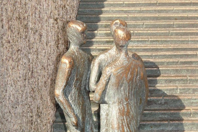Drei stilisierte menschliche Figuren aus Bronze stehen nebeneinander an einer Treppe.